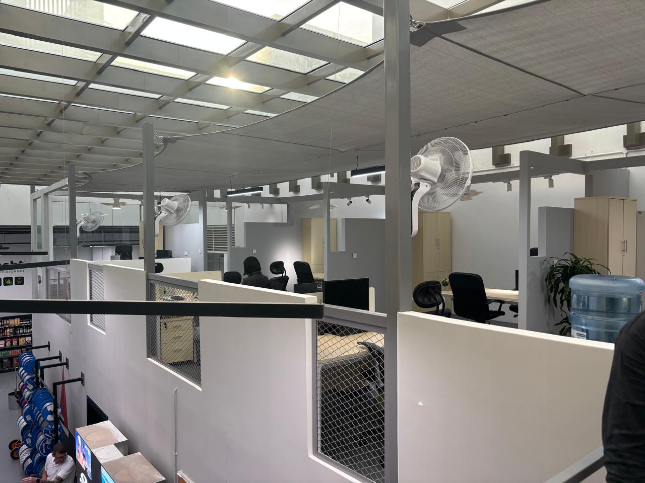 Mezzanine office with glass skylight ceiling, white cubicle partitions, mesh panels, fabric light canopy, and wall-mounted fans